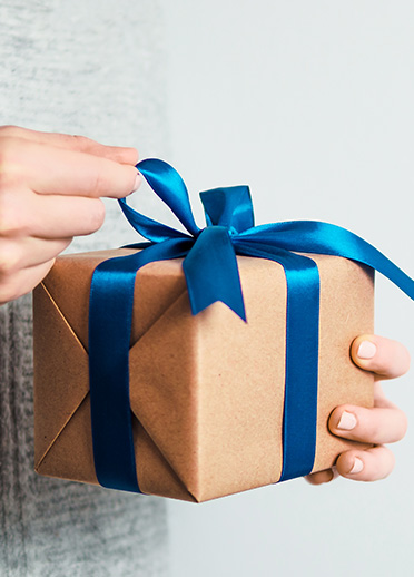 Die Abbildung zeigt eine Person, die ein Geschenk in braunem Papier mit einem blauen Satinband hält. Eine Hand zieht an der Schleife, um das Geschenk zu öffnen. 